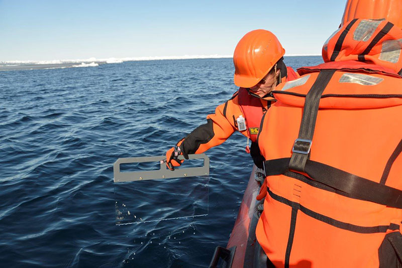 Samples of the surface film are taken with a glass plate during the Polarstern expedition PS106 2017 in the Arctic Ocean north of Spitsbergen. In order to rule out the influence of the icebreaker, the researchers travel to the open ocean in survival suits in a rubber dinghy. Photo: Marcel Köning, Christian-Albrechts-Universität zu Kiel